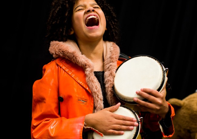 Een tiener met een oranje jas die bongo's vasthoudt en hardop zingt